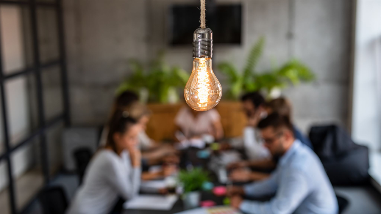 Equipo de trabajo en un salón de reuniones. En primer plano aparece una bombilla encendida - haciendo referencia a las nuevas ideas y la creatividad.